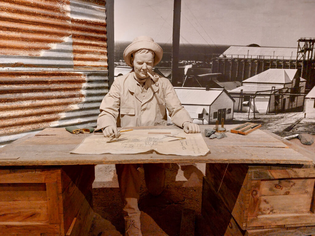 Likeness of Herbert Hoover sitting at a desk in a mine area looking over blueprints