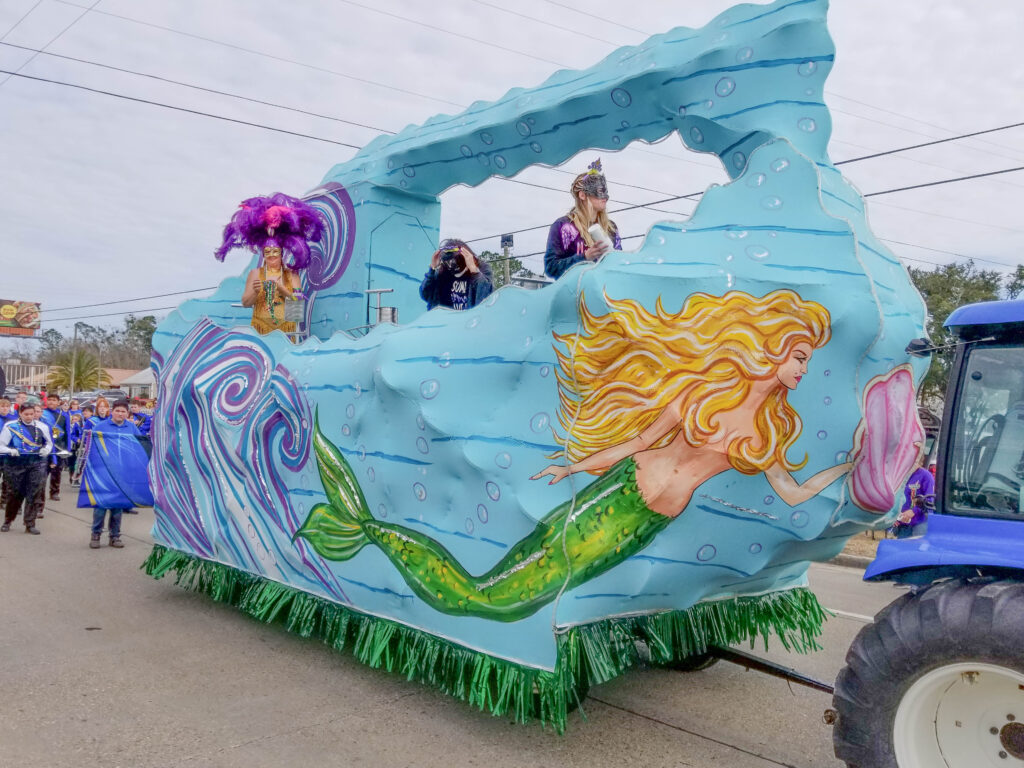 A float with a mermaid painted on it in the parade with three people wearing masks and costumes riding on it.