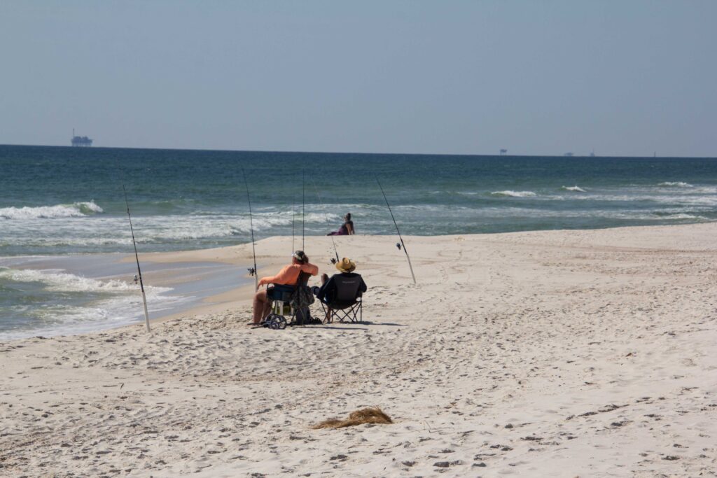 Two people on the beach, sitting on chairs and fishing.