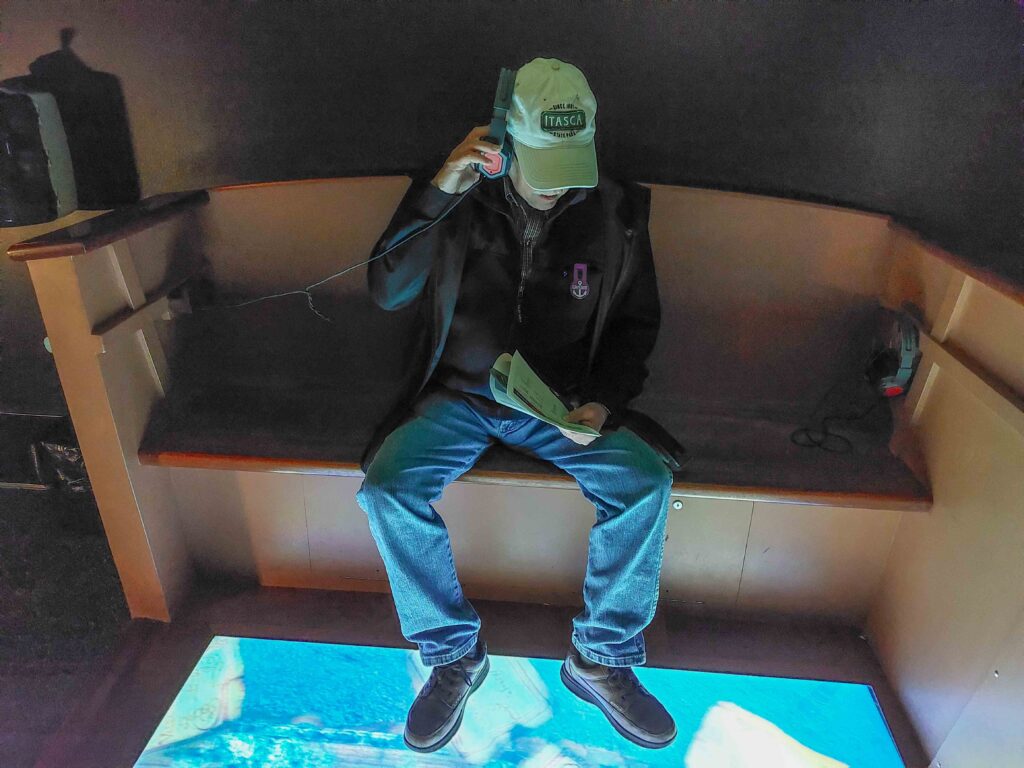 A man seated in a booth using a headset and looking at a lighted simulated sea beneath his feet
