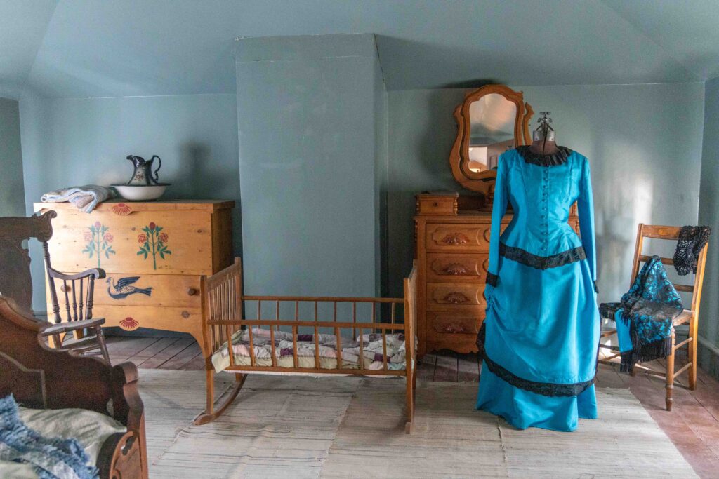 A bedroom in the Officers' Quarters. A blue 1800s woman's dress, baby cradle and a dresser.